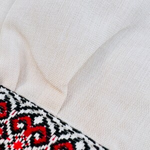 Op de afbeelding: Close-up van een beige stof met een geweven textuur. Een decoratieve rand met een geometrisch patroon in rode, zwarte en witte accenten accentueert de rand van de stof. De stof lijkt een kussen te zijn.