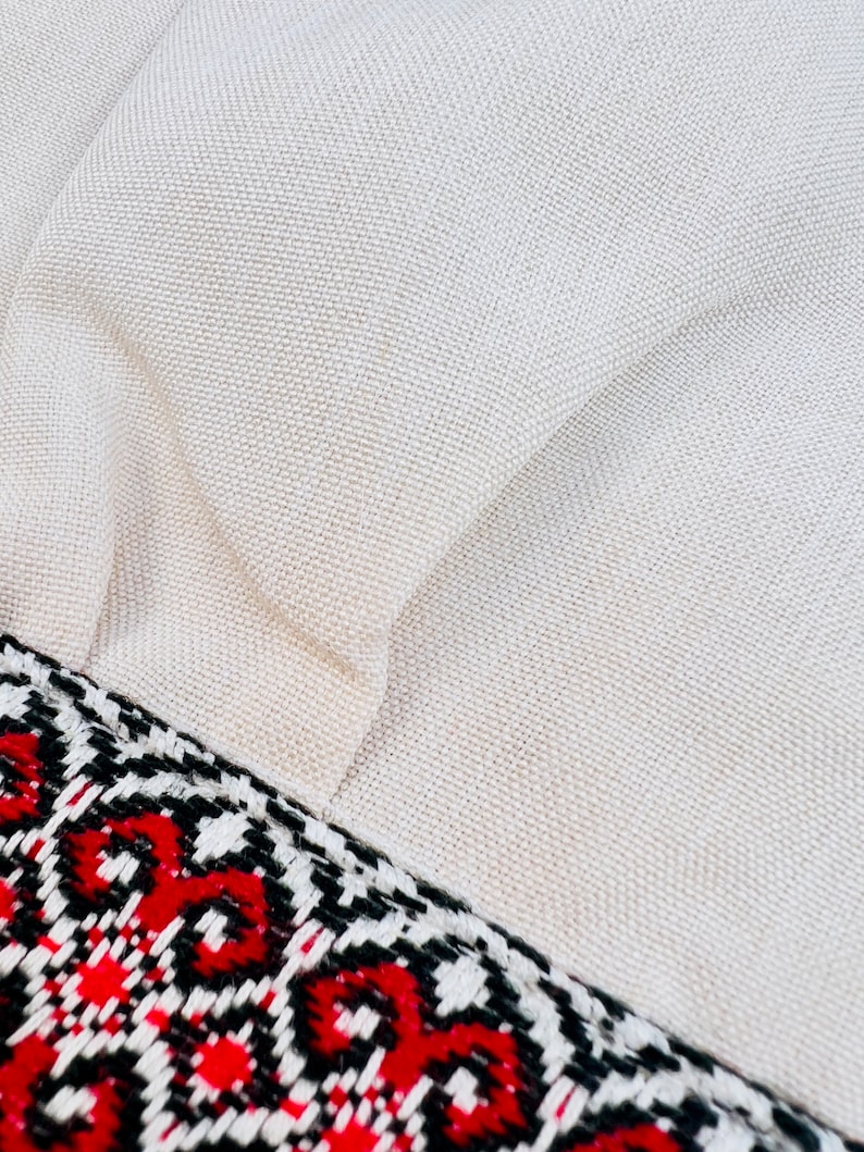 Op de afbeelding: Close-up van een beige stof met een geweven textuur. Een decoratieve rand met een geometrisch patroon in rode, zwarte en witte accenten accentueert de rand van de stof. De stof lijkt een kussen te zijn.