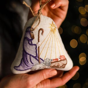 May include: Embroidered Christmas ornament in the shape of a bell. The ornament features a nativity scene with a shepherd in purple robes, a star, and baby Jesus. The ornament is made of a light-colored fabric and has a twine loop for hanging.