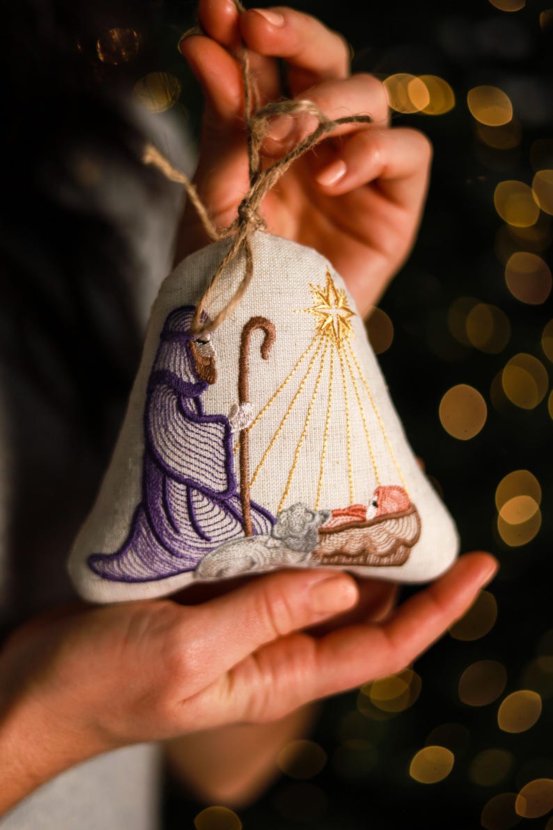 May include: Embroidered Christmas ornament in the shape of a bell. The ornament features a nativity scene with a shepherd in purple robes, a star, and baby Jesus. The ornament is made of a light-colored fabric and has a twine loop for hanging.