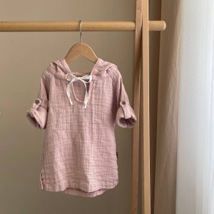 May include: A pale pink hooded shirt with a white ribbon tie at the neck. The shirt has a slightly loose fit and is made of a soft, textured fabric.
