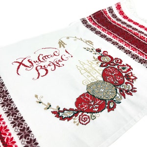 以下が含まれることがあります： 白い刺繍入り織物。縁には赤と茶色の幾何学模様が施されています。中央には赤、茶色、金色の花柄のデザインがあり、赤い糸でテキストが刺繍されています。この織物は装飾品である可能性があります。