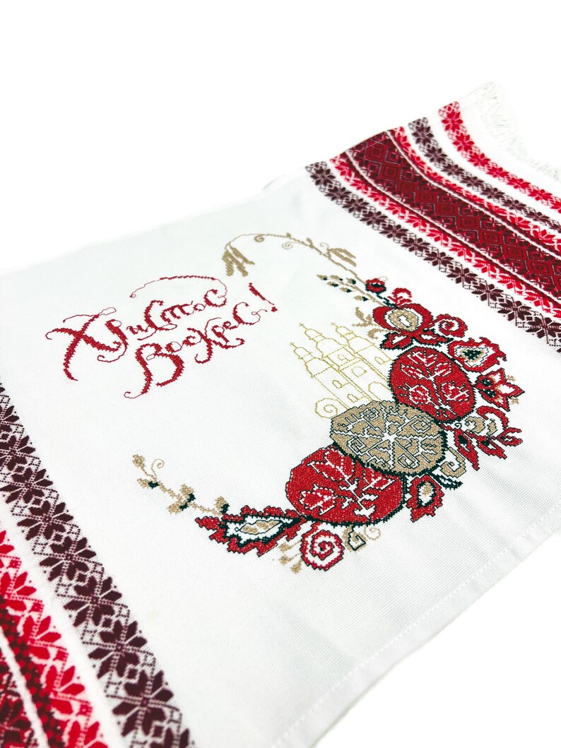 以下が含まれることがあります： 白い刺繍入り織物。縁には赤と茶色の幾何学模様が施されています。中央には赤、茶色、金色の花柄のデザインがあり、赤い糸でテキストが刺繍されています。この織物は装飾品である可能性があります。