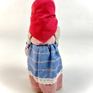 May include: A handmade doll wearing a red bonnet, a blue and white striped skirt, and a pink and white lace trim.