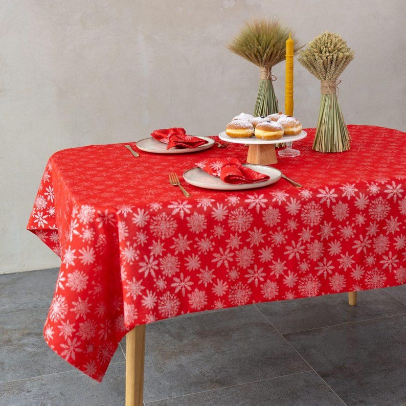 May include: A red tablecloth with a white snowflake pattern, set for a festive occasion. The table is set with plates, napkins, and cutlery. A cake stand holds donuts, and decorative wheat bundles and a candle complete the scene.