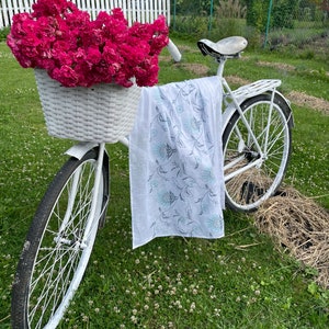 Puede incluir: Una bicicleta blanca con una cesta de mimbre llena de rosas rosas vibrantes. Una bufanda blanca con un estampado floral cuelga del manillar. La bicicleta est&aacute; sobre c&eacute;sped, con una valla blanca al fondo.