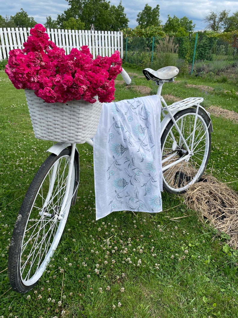 Puede incluir: Una bicicleta blanca con una cesta de mimbre llena de rosas rosas vibrantes. Una bufanda blanca con un estampado floral cuelga del manillar. La bicicleta est&aacute; sobre c&eacute;sped, con una valla blanca al fondo.