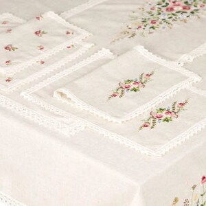 May include: Cream-colored linen tablecloth and matching napkins with floral embroidery. The tablecloth features a delicate lace trim and embroidered flowers along the edges and center. The napkins have embroidered floral designs.