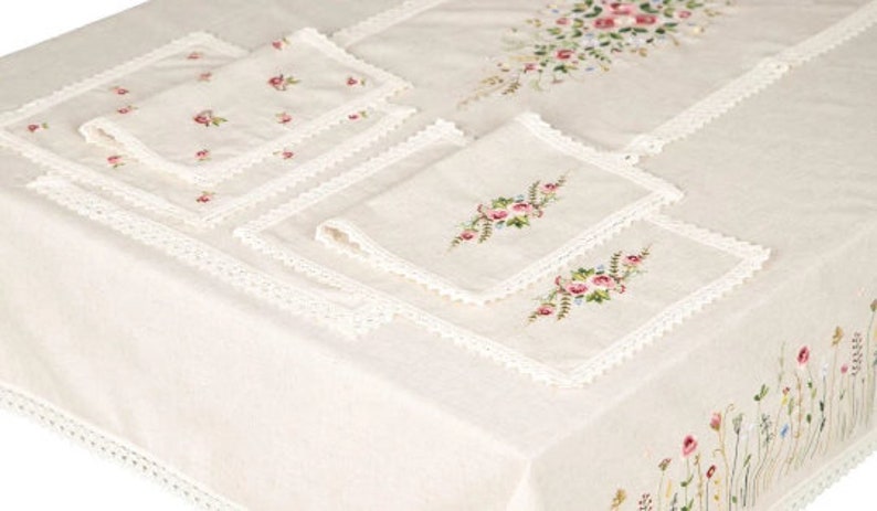 May include: Cream-colored linen tablecloth and matching napkins with floral embroidery. The tablecloth features a delicate lace trim and embroidered flowers along the edges and center. The napkins have embroidered floral designs.