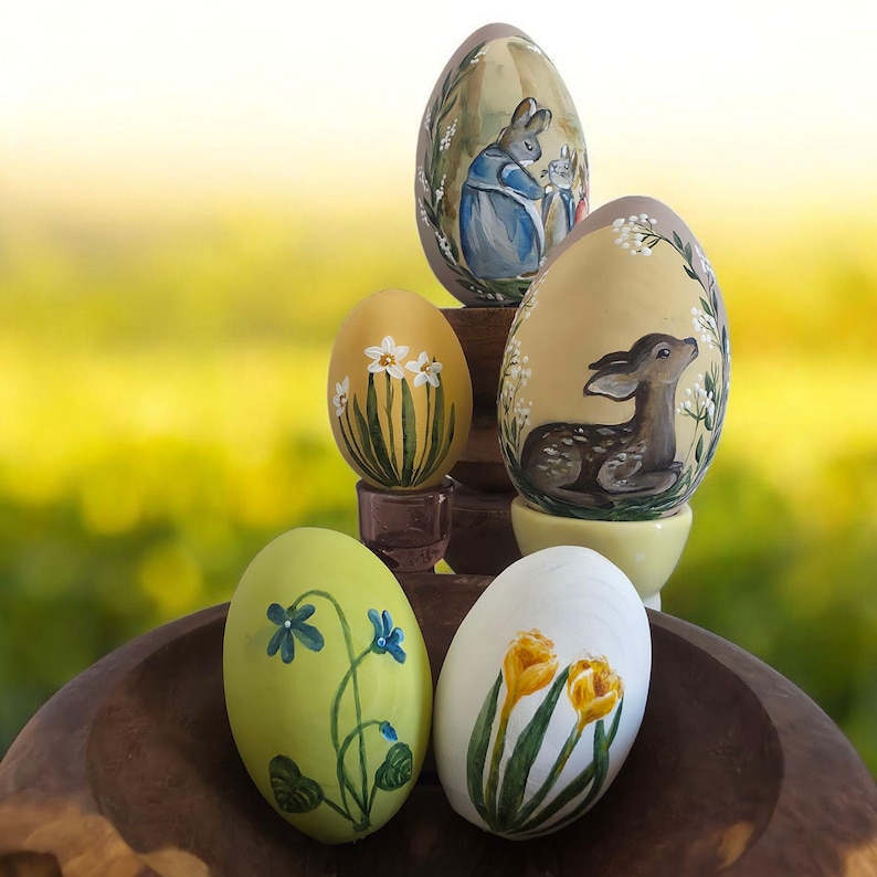 May include: A collection of hand-painted Easter eggs. The eggs feature various designs, including a bunny, a fawn, and floral patterns. The eggs are displayed on a wooden surface with a blurred green background.