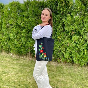 以下が含まれることがあります： 白い刺繍入りのブラウスと白いパンツを着用した女性が、刺繍の花が施された黒いトートバッグを持っています。バッグには、赤、黄、青の花のフラワーデザインが施されています。