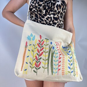 Puede incluir: Una bolsa de lona beige con un dise&ntilde;o floral bordado de colores. Las flores son en tonos de rojo, naranja, amarillo, verde, azul y rosa.