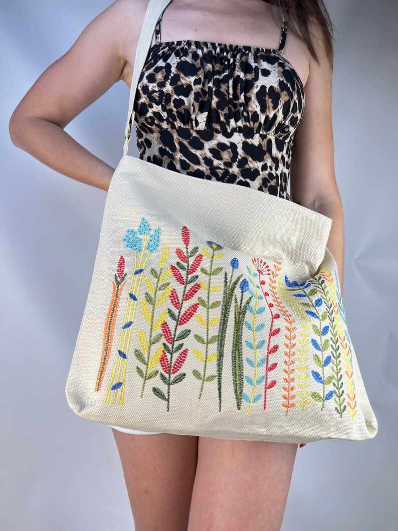 Puede incluir: Una bolsa de lona beige con un dise&ntilde;o floral bordado de colores. Las flores son en tonos de rojo, naranja, amarillo, verde, azul y rosa.
