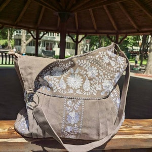 Peut inclure: Sac &agrave; bandouli&egrave;re marron avec broderies florales blanches et bleu clair. Le sac a un rabat et une longue bandouli&egrave;re. Fabriqu&eacute; en tissu robuste, il repose sur un banc en bois.