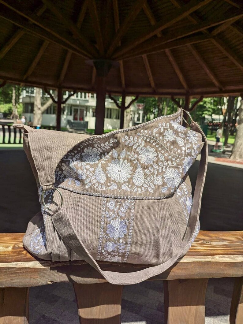 Peut inclure: Sac &agrave; bandouli&egrave;re marron avec broderies florales blanches et bleu clair. Le sac a un rabat et une longue bandouli&egrave;re. Fabriqu&eacute; en tissu robuste, il repose sur un banc en bois.