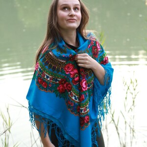 May include: A vibrant blue shawl with a floral pattern featuring red roses and green leaves. The shawl has a fringed edge and is worn over a black top. The background includes a body of water and greenery.
