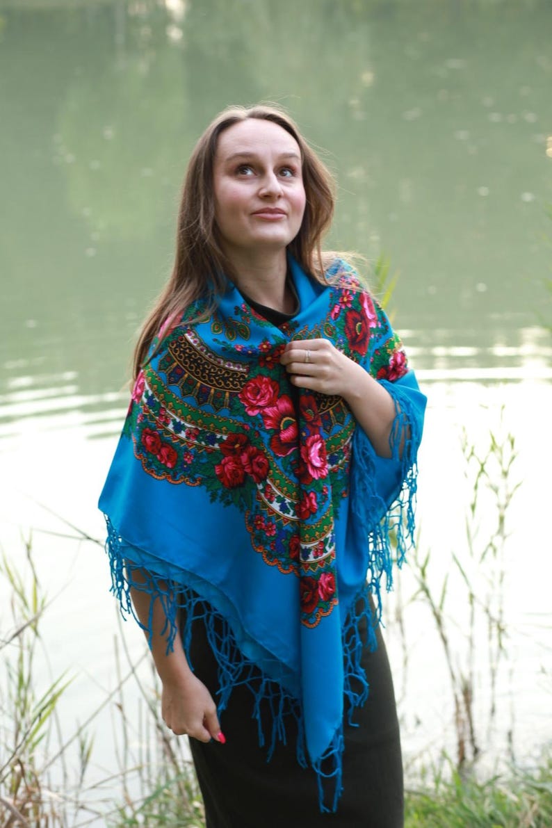 May include: A vibrant blue shawl with a floral pattern featuring red roses and green leaves. The shawl has a fringed edge and is worn over a black top. The background includes a body of water and greenery.
