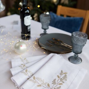 Puede incluir: Una mesa festiva con servilletas blancas bordadas, platos grises y copas de vino. Una botella de vino y un adorno navide&ntilde;o tambi&eacute;n est&aacute;n sobre la mesa, con un &aacute;rbol de Navidad borroso al fondo.