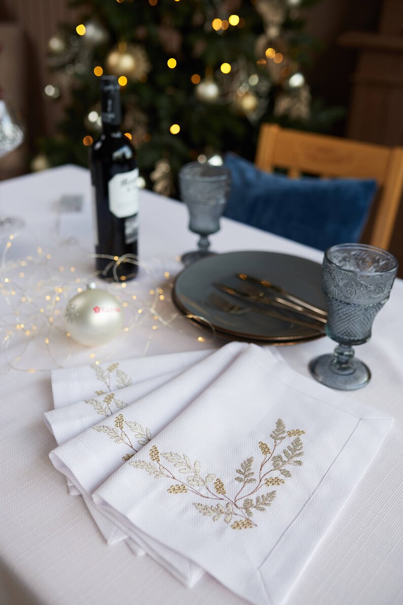 Puede incluir: Una mesa festiva con servilletas blancas bordadas, platos grises y copas de vino. Una botella de vino y un adorno navide&ntilde;o tambi&eacute;n est&aacute;n sobre la mesa, con un &aacute;rbol de Navidad borroso al fondo.