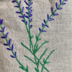 K&ouml;nnte beinhalten: Nahaufnahme von gestickten Lavendelzweigen auf Leinenstoff. Die Lavendelbl&uuml;ten sind violett und die Stiele gr&uuml;n. Der Stoff hat ein nat&uuml;rliches, strukturiertes Aussehen.