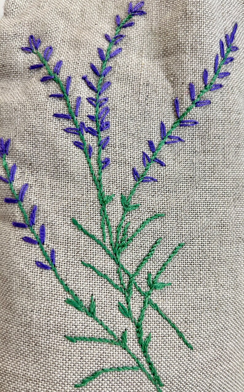 K&ouml;nnte beinhalten: Nahaufnahme von gestickten Lavendelzweigen auf Leinenstoff. Die Lavendelbl&uuml;ten sind violett und die Stiele gr&uuml;n. Der Stoff hat ein nat&uuml;rliches, strukturiertes Aussehen.