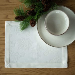 Op de afbeelding: Een witte rechthoekige placemat met een reli&euml;f bloemenontwerp, een bord en een kom. Het bord en de kom zijn lichtbeige met een bruine rand. Een takje dennen en dennenappels staan op de achtergrond. De items staan op een houten oppervlak.