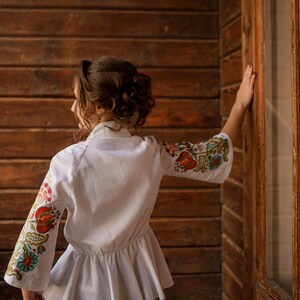 Op de afbeelding: Een wit linnen blouse met geborduurde bloemenmotieven op de mouwen. De blouse heeft een gerimpelde zoom en een getailleerd bovenlichaam.