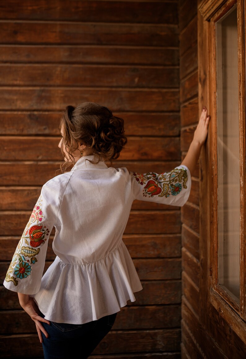 Op de afbeelding: Een wit linnen blouse met geborduurde bloemenmotieven op de mouwen. De blouse heeft een gerimpelde zoom en een getailleerd bovenlichaam.