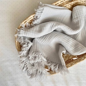 Peut inclure: Une couverture b&eacute;b&eacute; douce, grise et frang&eacute;e dans un panier tiss&eacute;. La couverture est faite d'un tissu l&eacute;ger et respirant.