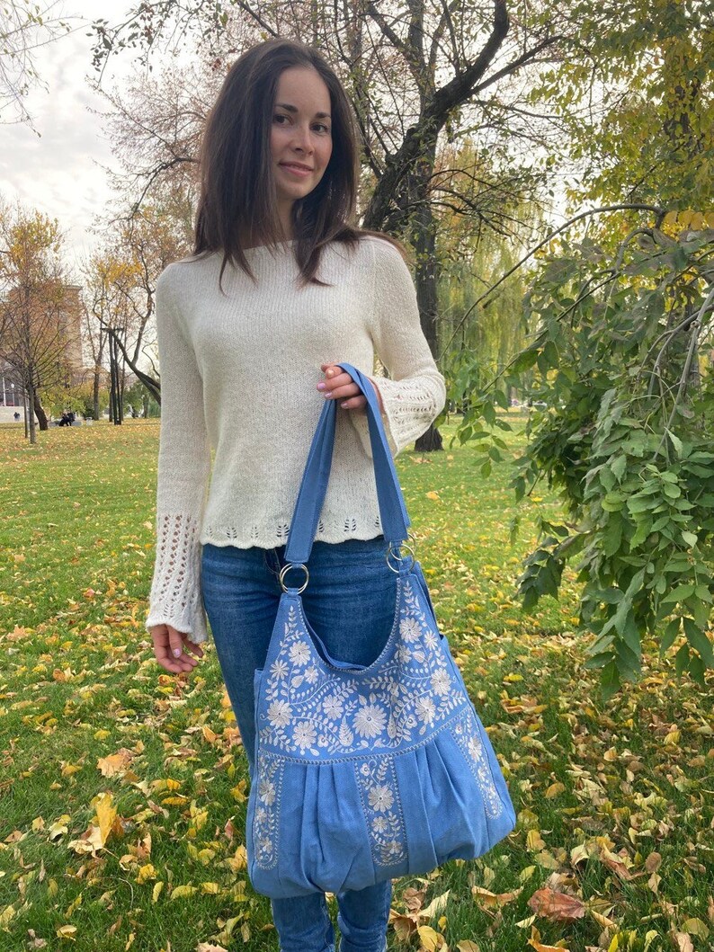 Peut inclure: Un sac cabas bleu avec des motifs floraux brod&eacute;s blancs. Le sac a un haut large et ouvert et deux longues anses.