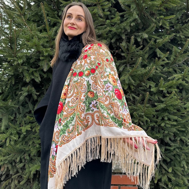 May include: A white shawl with a floral pattern in shades of red, green, and gold. The shawl has a fringe border and is worn over a black coat.
