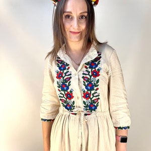 Pode incluir: Uma mulher vestindo um vestido de linho branco com bordados florais coloridos. Ela est&aacute; usando uma coroa de flores com flores vermelhas, amarelas e azuis.