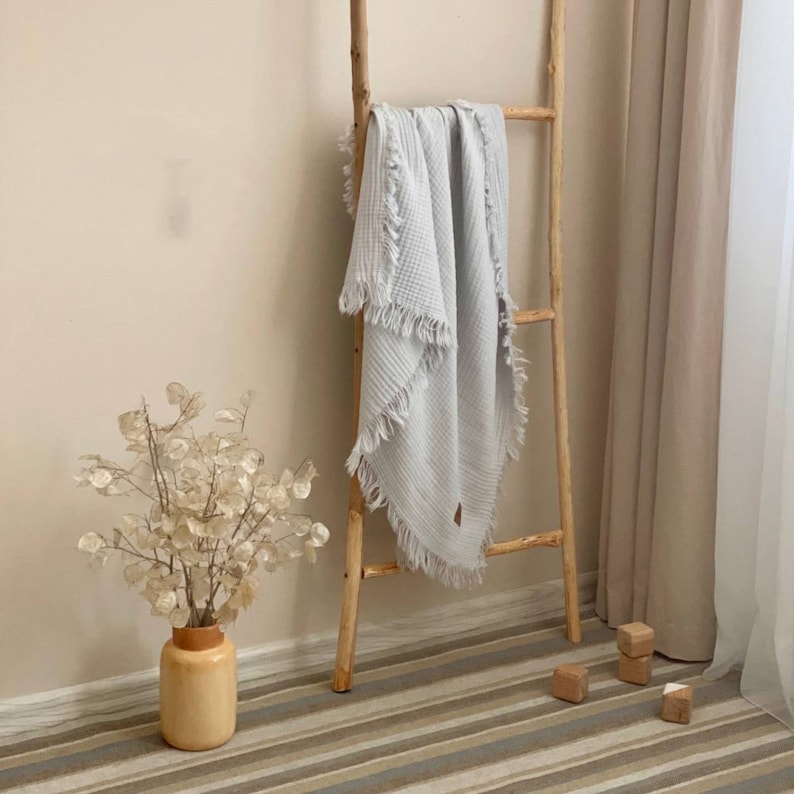 Peut inclure: Une couverture en tissu gaufr&eacute; gris clair avec des bords frang&eacute;s, drap&eacute; sur une &eacute;chelle en bois. La couverture est suspendue &agrave; une &eacute;chelle en bois contre un mur beige clair.