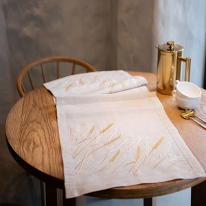 Op de afbeelding: Een beige linnen tafelloper met geborduurde tarwearen in goud- en cr&egrave;metinten. De loper ligt op een ronde houten tafel, met een gouden koffiepers, witte kopjes en gouden lepels op de achtergrond. De tafel is gedekt voor een maaltijd.