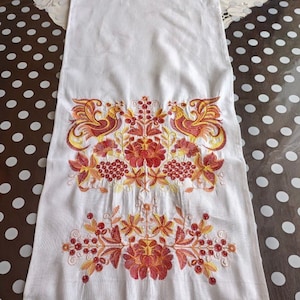 Peut inclure: Chemin de table en lin blanc avec broderies complexes rouges, oranges et jaunes. Le motif pr&eacute;sente des oiseaux stylis&eacute;s et des motifs floraux. Le bord inf&eacute;rieur est orn&eacute; d'une bordure en dentelle rouge, ajoutant une touche d&eacute;corative &agrave; ce d&eacute;cor &eacute;l&eacute;gant.