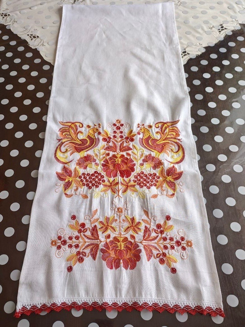 Peut inclure: Chemin de table en lin blanc avec broderies complexes rouges, oranges et jaunes. Le motif pr&eacute;sente des oiseaux stylis&eacute;s et des motifs floraux. Le bord inf&eacute;rieur est orn&eacute; d'une bordure en dentelle rouge, ajoutant une touche d&eacute;corative &agrave; ce d&eacute;cor &eacute;l&eacute;gant.
