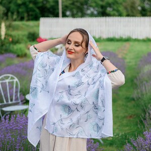 Puede incluir: Un chal de lino blanco con un estampado floral en tonos azules y grises. El chal est&aacute; drapeado sobre la cabeza y los hombros. La persona lleva un vestido beige y est&aacute; de pie en un campo de lavanda.