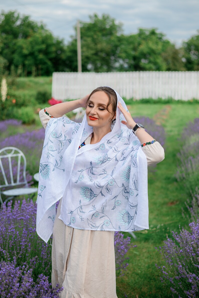 Puede incluir: Un chal de lino blanco con un estampado floral en tonos azules y grises. El chal est&aacute; drapeado sobre la cabeza y los hombros. La persona lleva un vestido beige y est&aacute; de pie en un campo de lavanda.