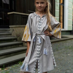 K&ouml;nnte beinhalten: Graues Leinenkleid mit gestickten Details und passendem G&uuml;rtel. Das Kleid hat gelbe Weizen&auml;hren auf den &Auml;rmeln und wei&szlig;e und blaue Stickereien an der Vorderseite und am Saum. Das Kleid hat einen knielangen Saum und ist mit Quasten verziert.
