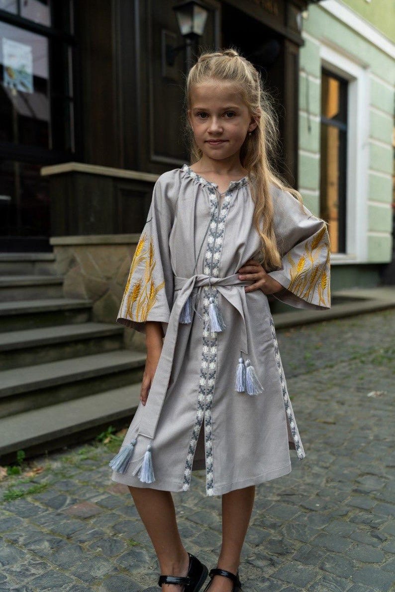 K&ouml;nnte beinhalten: Graues Leinenkleid mit gestickten Details und passendem G&uuml;rtel. Das Kleid hat gelbe Weizen&auml;hren auf den &Auml;rmeln und wei&szlig;e und blaue Stickereien an der Vorderseite und am Saum. Das Kleid hat einen knielangen Saum und ist mit Quasten verziert.
