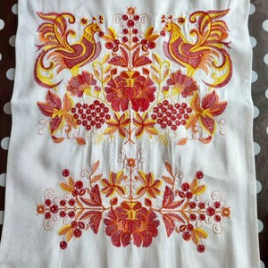 May include: White linen tea towel with red and yellow embroidered birds and floral designs. The towel has a red crocheted trim.