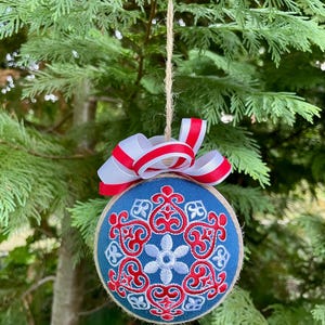K&ouml;nnte beinhalten: Ein blauer und wei&szlig;er Weihnachtsbaumschmuck mit einem rot-wei&szlig;en Band. Der Schmuck ist mit einem Schneeflockenmuster verziert.