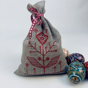 K&ouml;nnte beinhalten: Eine kleine graue Leinentasche mit einem roten gestickten Blumendesign und einem rot-wei&szlig; gepunkteten Band. Die Tasche ist gef&uuml;llt mit bunten, handbemalten Ostereiern.