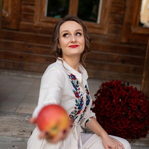 May include: A woman wearing a cream-colored dress with floral embroidery, holding a red apple. The dress features colorful floral patterns around the collar and cuffs. A large bouquet of red flowers is in the background.