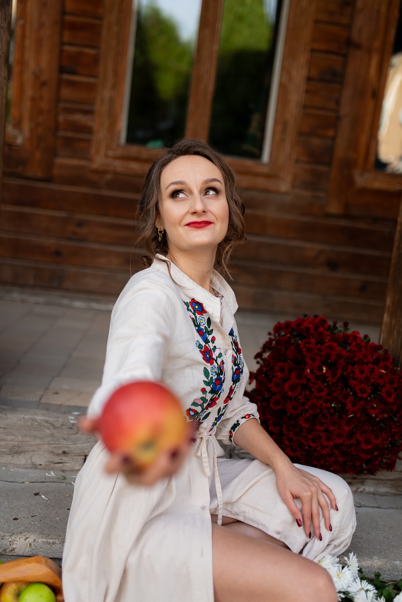 May include: A woman wearing a cream-colored dress with floral embroidery, holding a red apple. The dress features colorful floral patterns around the collar and cuffs. A large bouquet of red flowers is in the background.