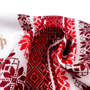 Puede incluir: Tela blanca con un detallado patr&oacute;n bordado en rojo y burdeos. El dise&ntilde;o presenta formas geom&eacute;tricas y motivos florales. La tela est&aacute; doblada, mostrando los intrincados detalles del bordado. El tema general es el arte textil tradicional.