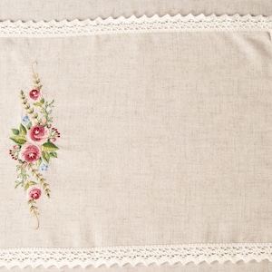 May include: A white linen placemat with a delicate lace border and a floral embroidery design featuring pink and red roses and green leaves.