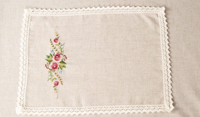 May include: A white linen placemat with a delicate lace border and a floral embroidery design featuring pink and red roses and green leaves.