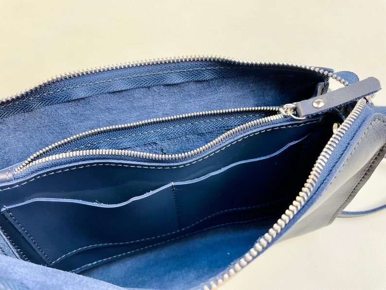 May include: A navy blue leather bag with two zippered compartments and a pocket inside. The bag is open and the interior lining is visible.