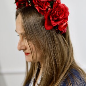 K&ouml;nnte beinhalten: Ein Blumenkranz mit gro&szlig;en, tiefroten Rosen. Der Kopfschmuck besteht aus realistisch aussehenden Rosen und kleinen roten Beeren. Die Person tr&auml;gt ein dunkelblaues Oberteil. Die roten Rosen sind ein Blickfang.
